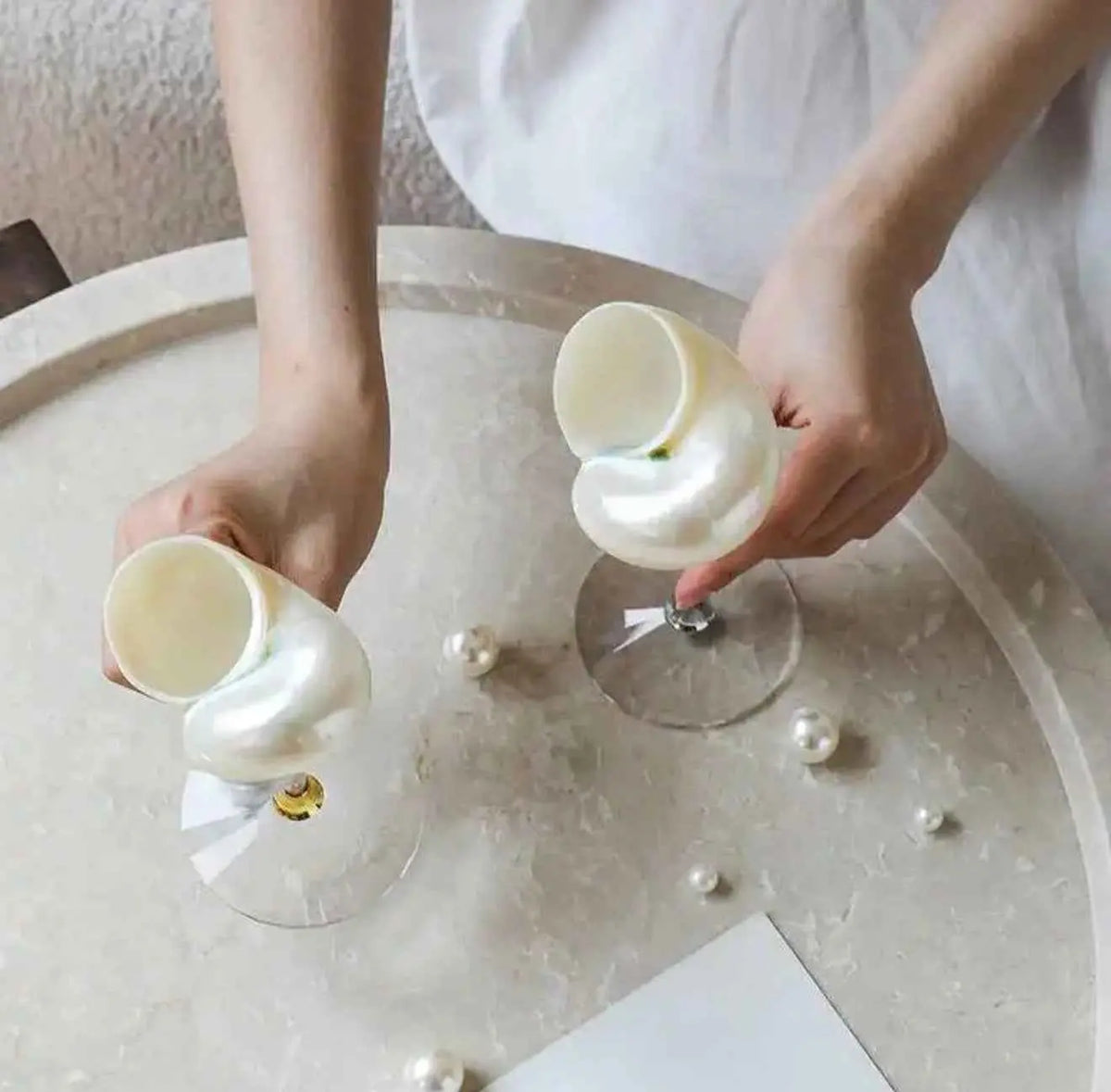Hands holding two white conch shell goblets over a round table